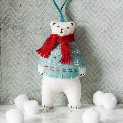 White teddy bear wearing a red scarf and patterned sweater against a chevron-patterned wall. Corinne Lapierre Polar Bear Felt Craft Kit – complete wool felt sewing kit with materials and instructions to hand-stitch a polar bear decoration