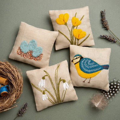 Four embroidered pillows with floral and bird designs on a green  Corinne Lapierre Linen Lavender Bag Spring Garden Craft Kit – complete sewing and embroidery kit with linen fabric, lavender, and materials to hand-stitch a scented bagbackground