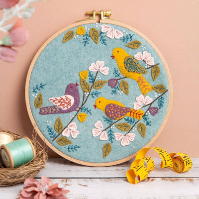 Embroidery hoop with colorful birds and flowers on a wooden surface. Corinne Lapierre Dawn Chorus Appliqué Hoop Craft Kit – complete embroidery and appliqué sewing kit with hoop, fabric, felt and materials to hand-stitch a bird-themed design