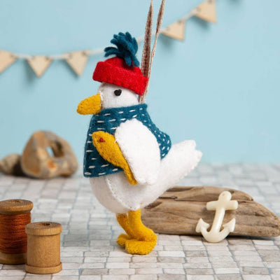 Toy duck wearing a red hat and blue sweater on a textured surface with driftwood and an anchor. Corinne Lapierre Sam the Seagull Felt Craft Kit – complete wool felt sewing kit with materials and instructions to hand-stitch Sam the Seagull decoration