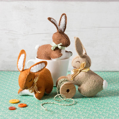 Three plush bunny toys with buttons and a spool of thread on a green  Corinne Lapierre Bunny Felt Craft Kit – complete wool felt sewing kit with materials and instructions to hand-stitch a bunny decorationsurface.