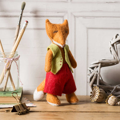 Stuffed fox toy wearing a green vest and red outfit on a wooden surface with decorative items. Corinne Lapierre Fergus Fox Felt Craft Kit – complete wool felt sewing kit with materials and instructions to hand-stitch a woodland fox decoration