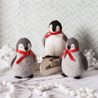 Three felt penguins with red polka dot bows on a marble surface with decorative balls and a chevron-patterned wall. Baby Penguins Felt Craft Kit – Complete DIY sewing kit with felt, thread, needle, stuffing, and instructions to make two adorable penguin decorations
