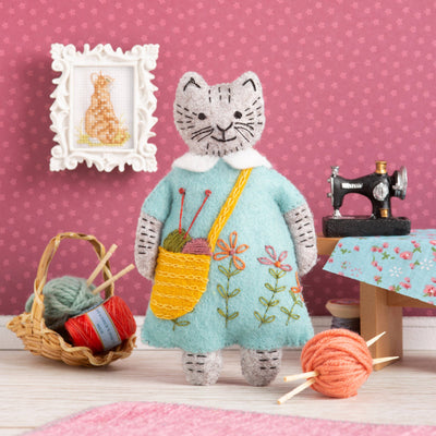 Cat-shaped plush toy in a room with sewing equipment and yarn on a pink polka dot wall background Mrs Cat Loves Knitting Felt Craft Mini Kit – Complete DIY sewing kit with felt, thread, needle, stuffing, and instructions to make a cat decoration