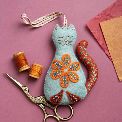 Embroidered cat decoration with floral patterns on a pink background, accompanied by sewing tools. Folk Cat Felt Craft Mini Kit – Complete DIY sewing kit with felt, thread, needle, stuffing, and instructions to make a folk-style cat decoration