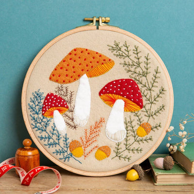 Embroidery hoop with colorful mushroom design on a wooden surface against a blue wall. Corinne Lapierre Toadstool Appliqué Hoop Craft Kit – complete appliqué and embroidery sewing kit with hoop, fabric, felt and materials to hand-stitch a woodland toadstool design