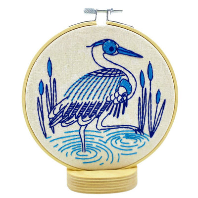 Embroidery hoop with a blue heron design on beige fabric. Hook, Line & Tinker Blue Heron Embroidery Kit – DIY embroidery kit featuring a modern folk-style heron design with fabric, hoop, floss, needle, and instructions