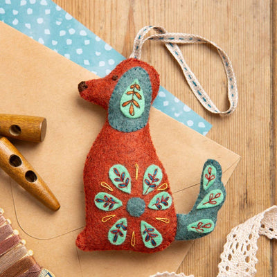 Decorative felt dog ornament with floral patterns on a wooden surface with craft materials. Corinne Lapierre Folk Dog Felt Craft Kit – complete wool felt sewing kit with materials and instructions to hand-stitch a folk-inspired dog decoration