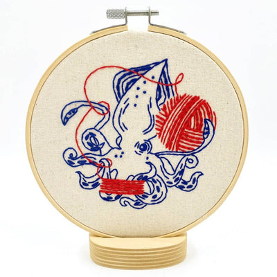 Embroidery hoop with a squid and yarn ball design on beige fabric.Hook, Line & Tinker Squid Balling Yarn Complete Embroidery Kit – DIY folk-style embroidery kit featuring a squid balling yarn, with hoop, printed fabric, floss, needle & eco-friendly packaging